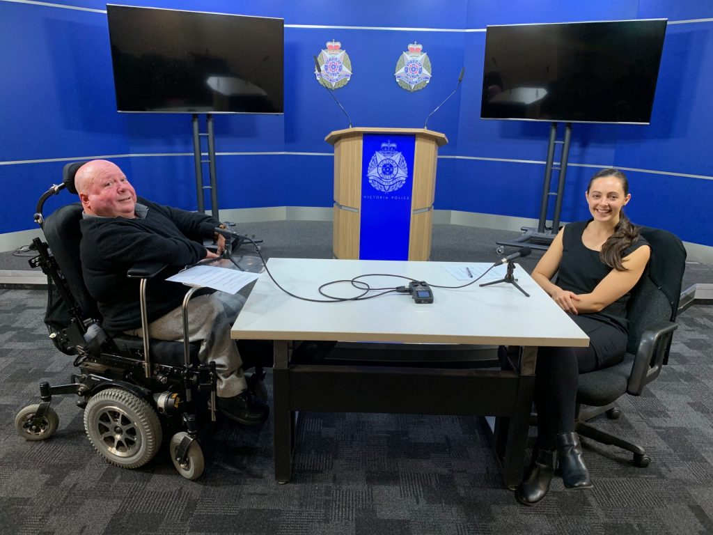 Man in wheelchair sitting at a desk across from woman. both are smiling at a camera